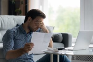 Stressed young man, covering eyes, holding document. Facing denial of claim by insurance company.