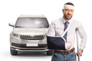 Driver with crutch post-car accident, standing by vehicle with shattered windshield on white background.