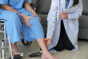 Compassionate female doctor attends to overworked male patient in a wheelchair with knee and ankle pain at the hospital, receiving necessary medical care.