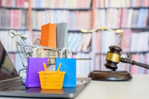 Representation of consumer rights and protection in business law. A laptop displays a shopping basket, shopping bags, and a shopping cart, accompanied by a judge's gavel symbolizing justice, a balanced scale indicating fairness, and a bookshelf in the background.