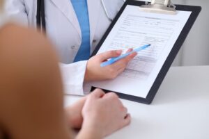 Close-up image of a doctor and patient's hands, with the physician gesturing towards details on a medical history form attached to a clipboard.