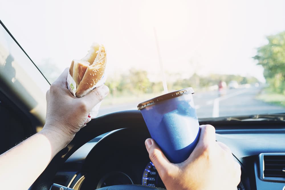 Man dangerously eating a hot dog and drinking a cold beverage while driving a car.